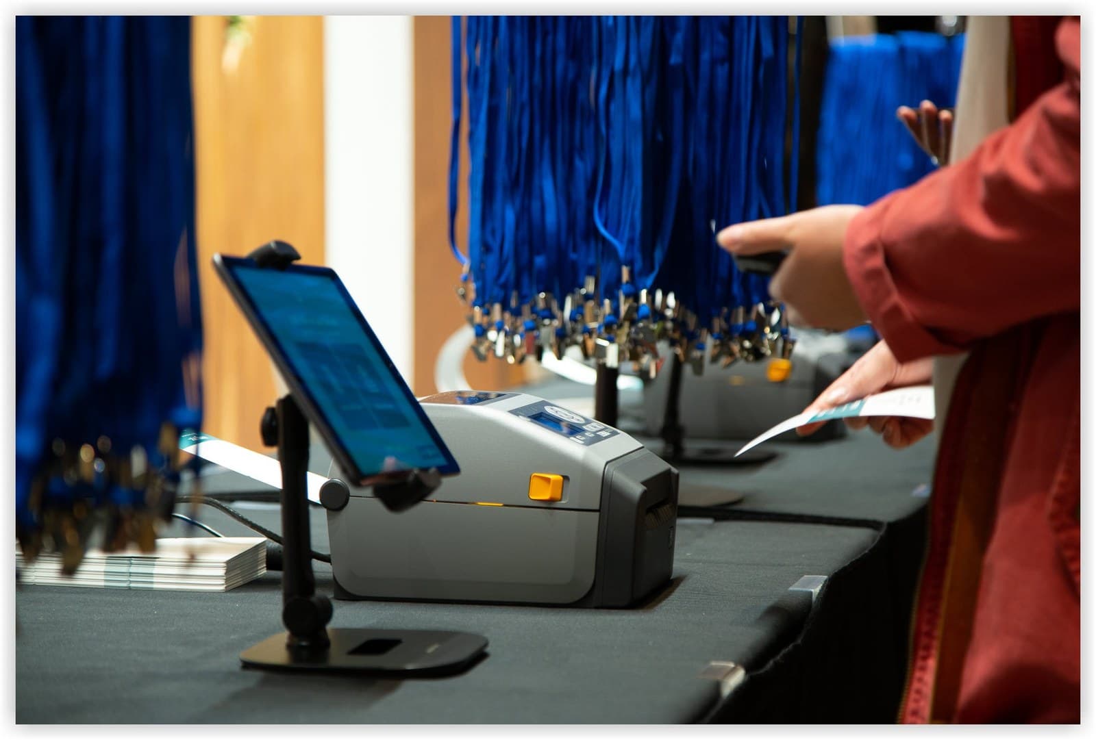 An iPad on a stand next to a badge printer, with blue lanyards hanging above